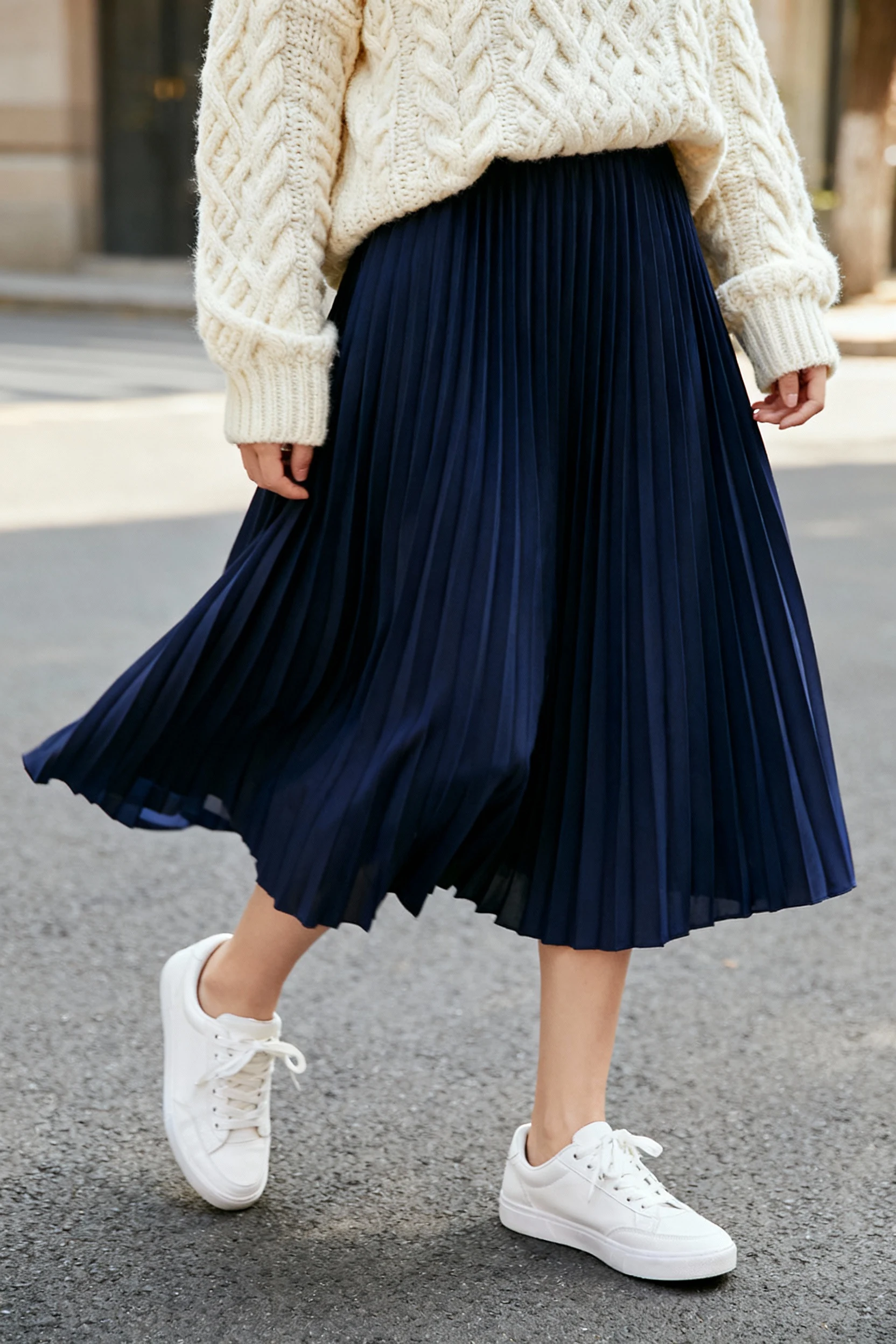 closeup female cream chunky sweater, navy pleated midi, white sneakers