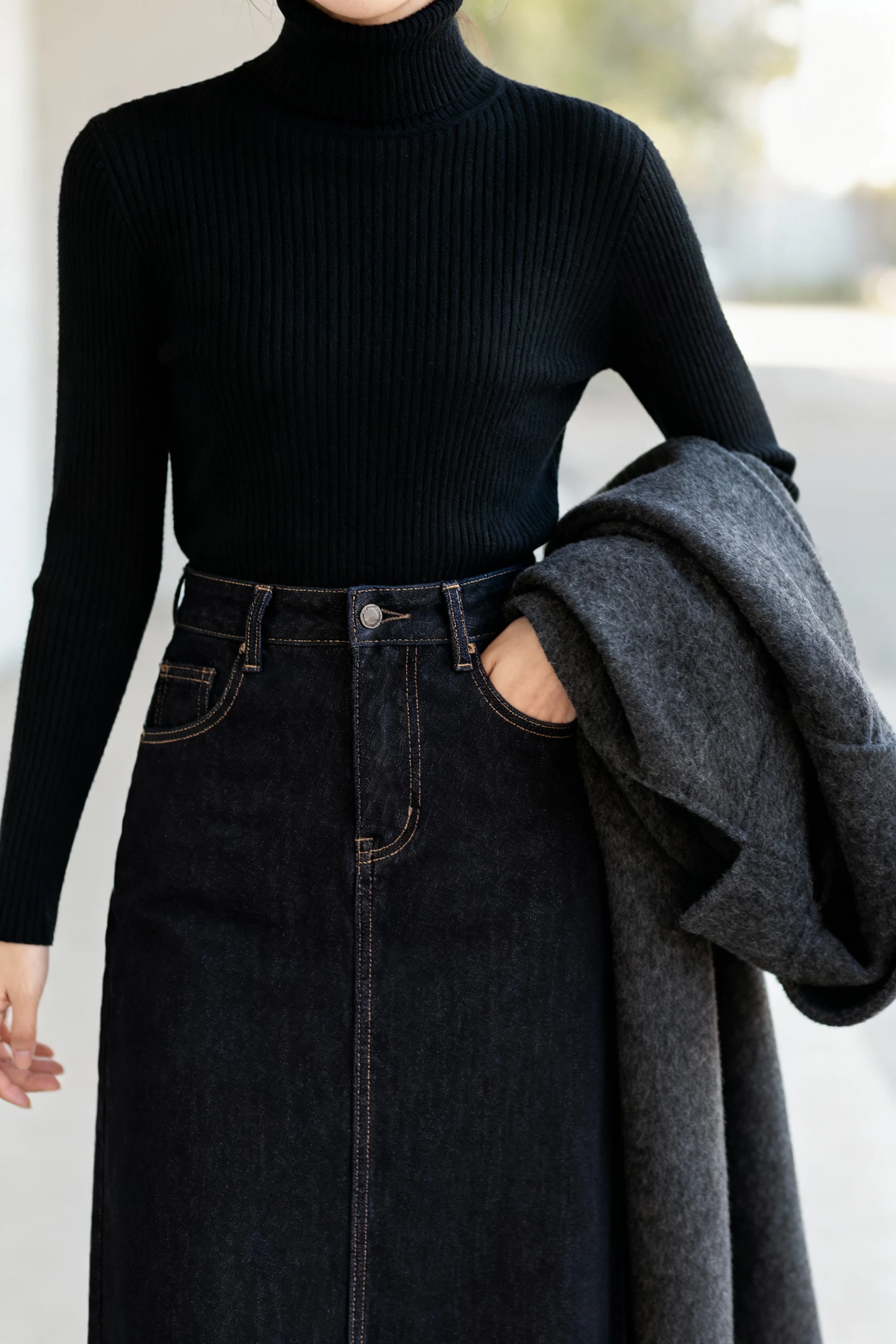 closeup of ribbed black turtleneck tucked into midi dark denim skirt with wool coat draped over arm