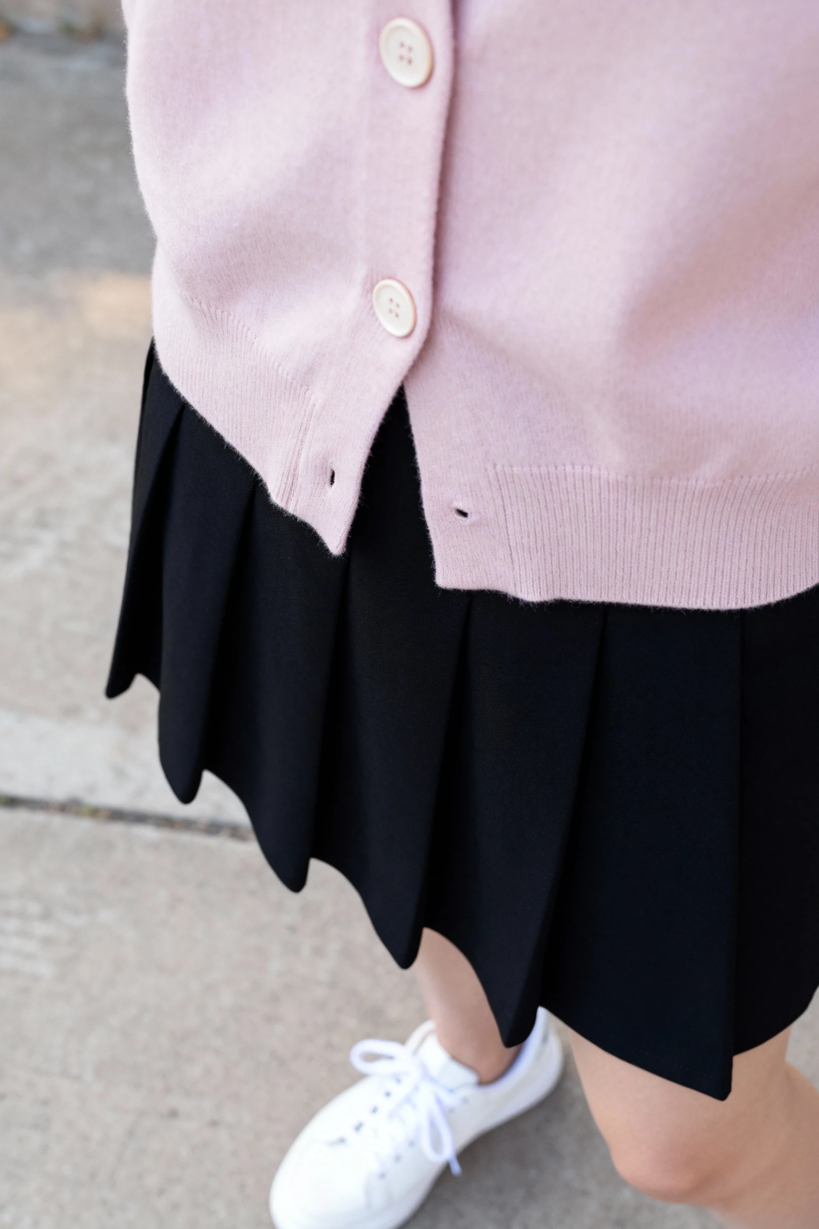 closeup of pastel cropped cardigan buttons over black mini skirt with white sneakers on concrete pavement